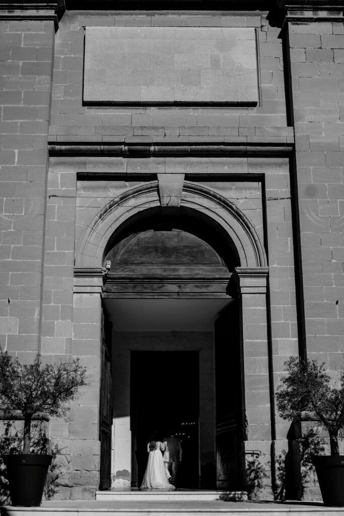 wedding photographer provence A bride and groom stand at the entrance of a large stone building with tall arched doors, captured in black and white. Sunlight highlights the couple as they enter the shadowed doorway of aix en provence main church