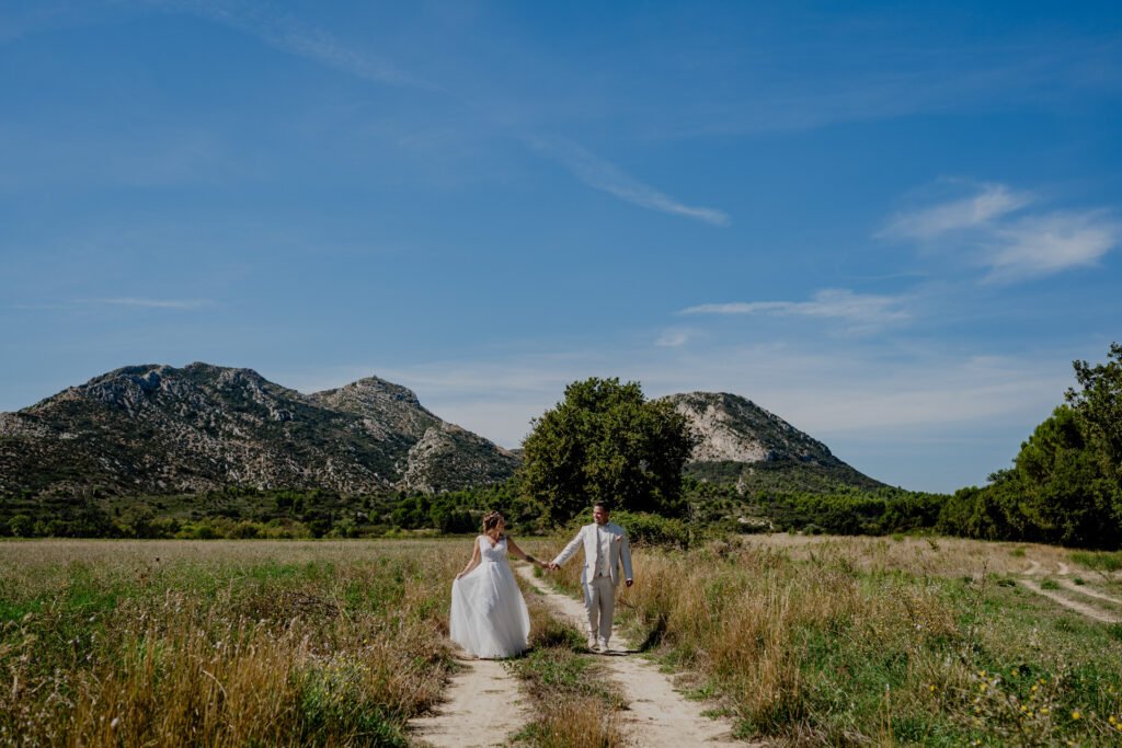 wedding photographer provence A bride and groom hold hands while walking down a dirt path in a grassy field, with mountains and trees in the background under a clear blue sky. in aix en provence close to lavander fields